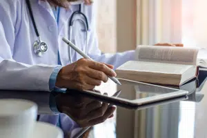 A doctor writing something on a tablet with a book open next to it.