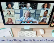 A desk with open notebooks, a keyboard, a white mouse, and a computer screen with a group therapy session taking place.