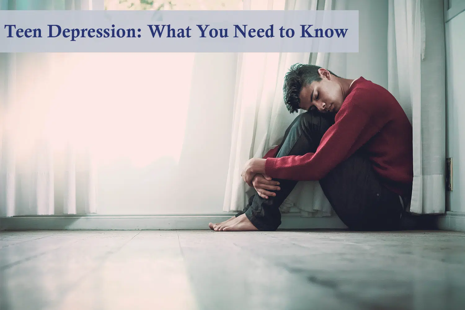 Boy in red sweater with teen depression sitting barefoot on the floor next to a window with gauzy curtains.